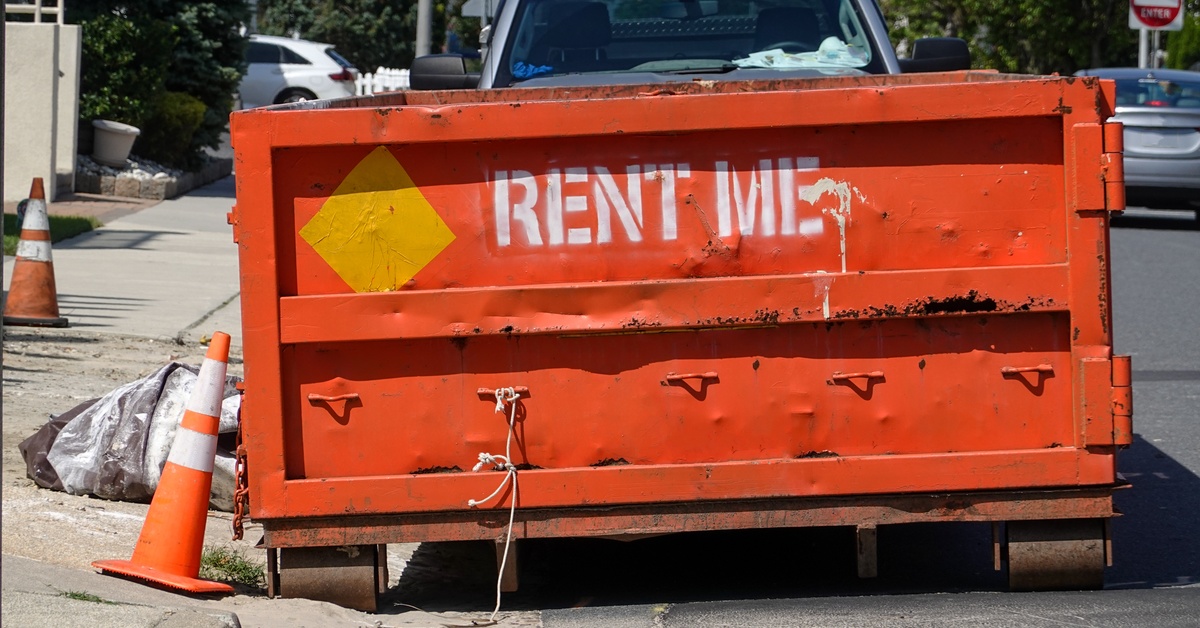 A close-up of an orange dumpster sitting on a curb. It says "RENT ME" in white spray-painted letters.