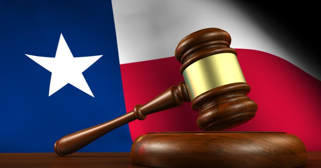 A close-up of a gavel on a wooden table with a Texas state flag in the background. The gavel sits upright.