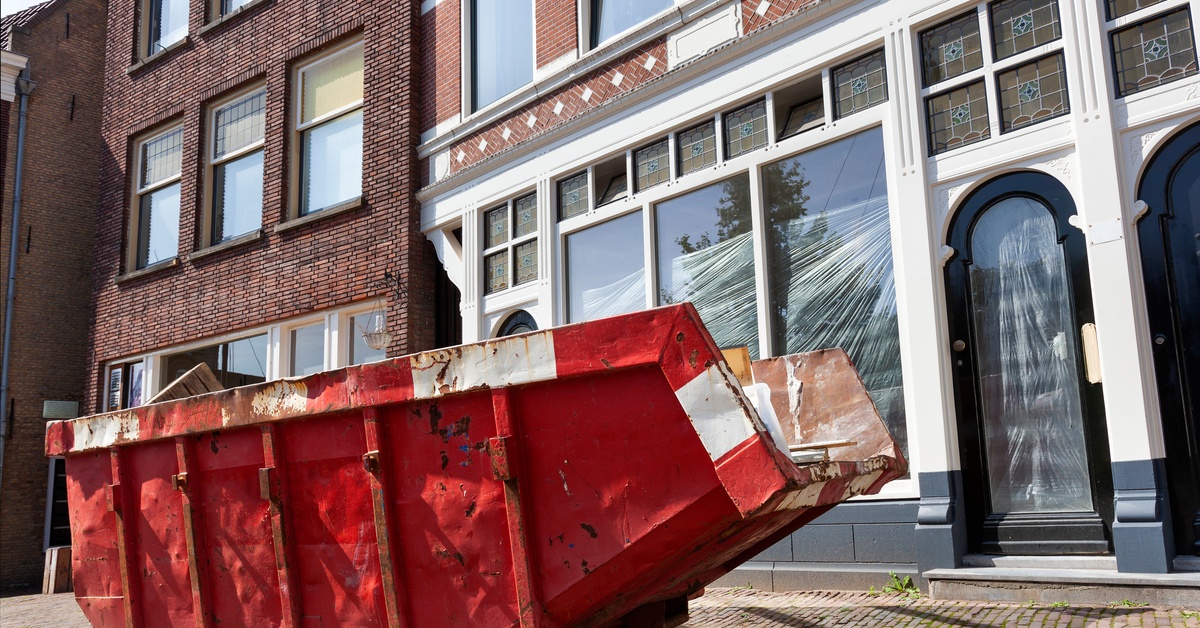 A red dumpster sits in front of a business storefront and apartment building. Items slightly poke out of the dumpster.