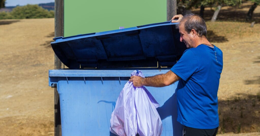 A man throws trash bags into a light-blue outdoor dumpster. The man wears a dark blue shirt and black pants.