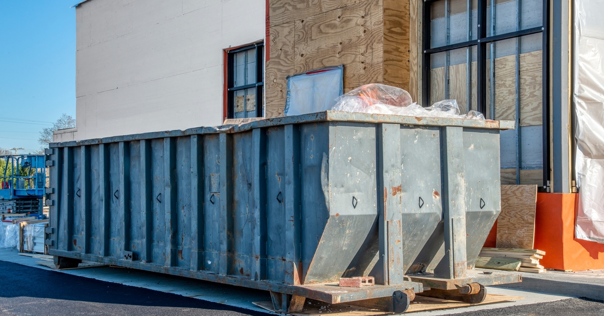 A large dumpster is placed next to a building. The building itself is unfinished and has wood covering various parts of the exterior.
