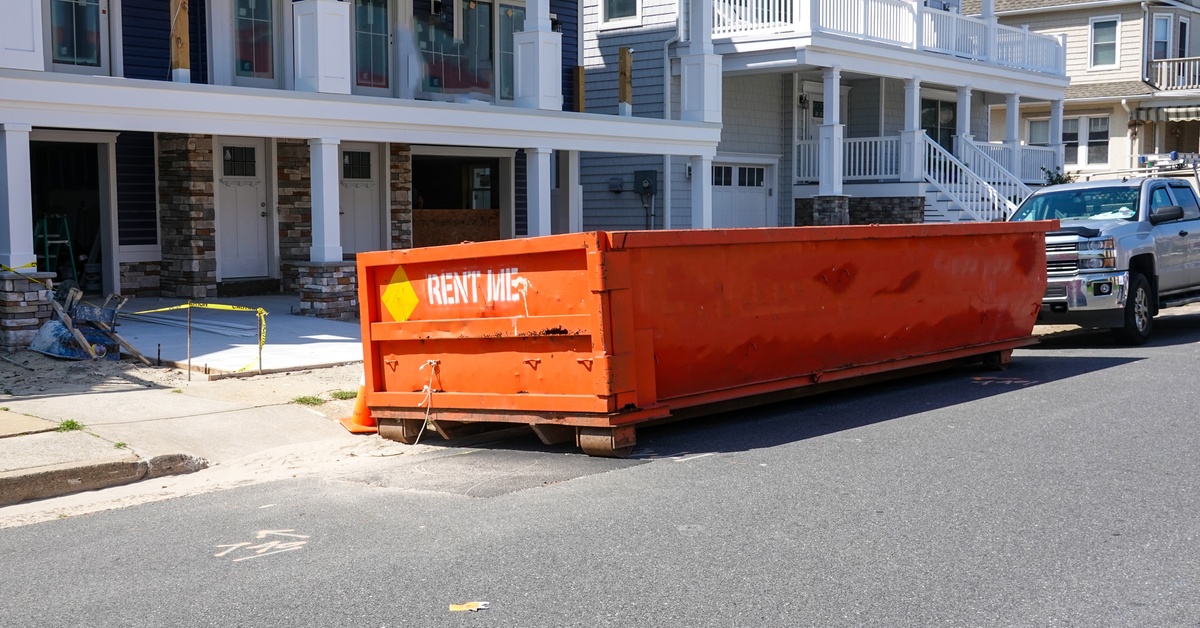 A large orange dumpster with "RENT ME" written on the side is placed on the street in front of an apartment complex.